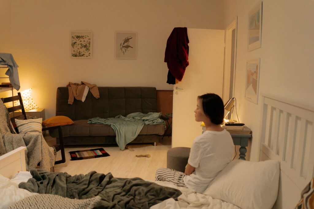 realistic-messy-bedroom A woman sits on a bed in a dimly lit, cluttered bedroom with clothes, blankets, and personal items scattered around.