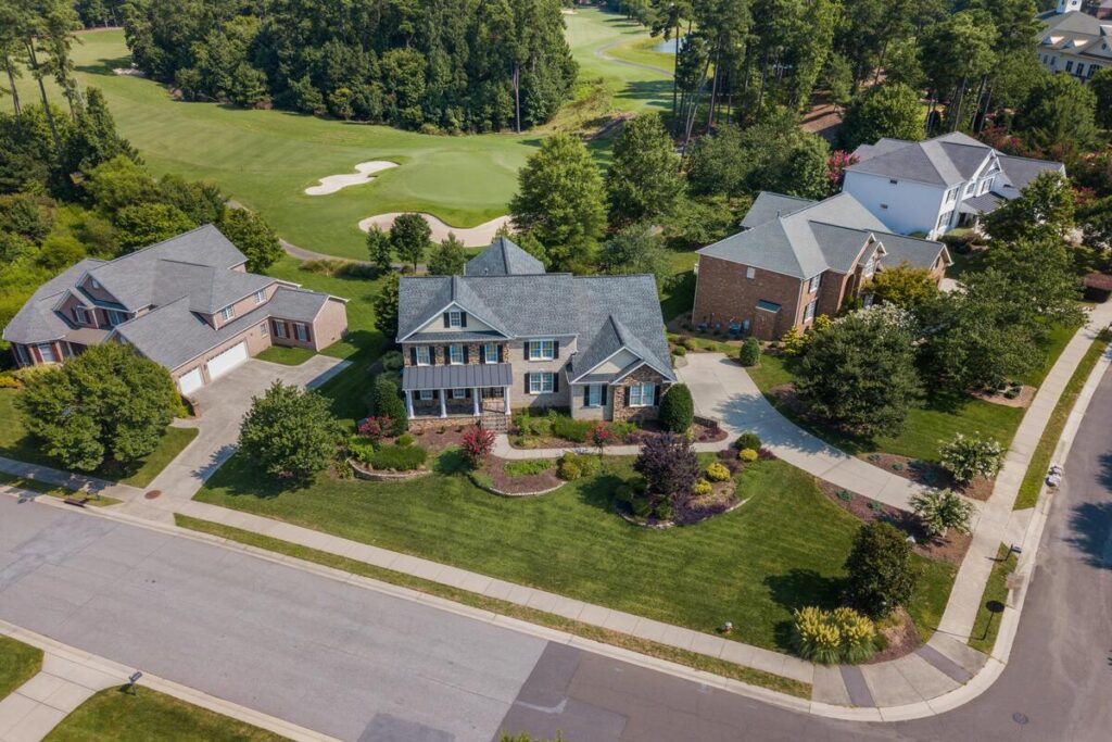 Aerial view of a corner lot home with a wide, well-kept front lawn, layered shrubs and trees along the street edges, and a clear driveway and entry path.